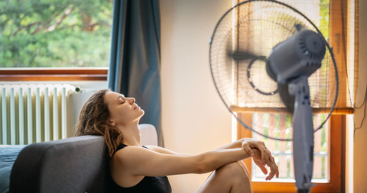 A woman is sitting in front of a fan, struggling with the heat because she did not properly maintain her AC with our spring HVAC maintenance checklist.